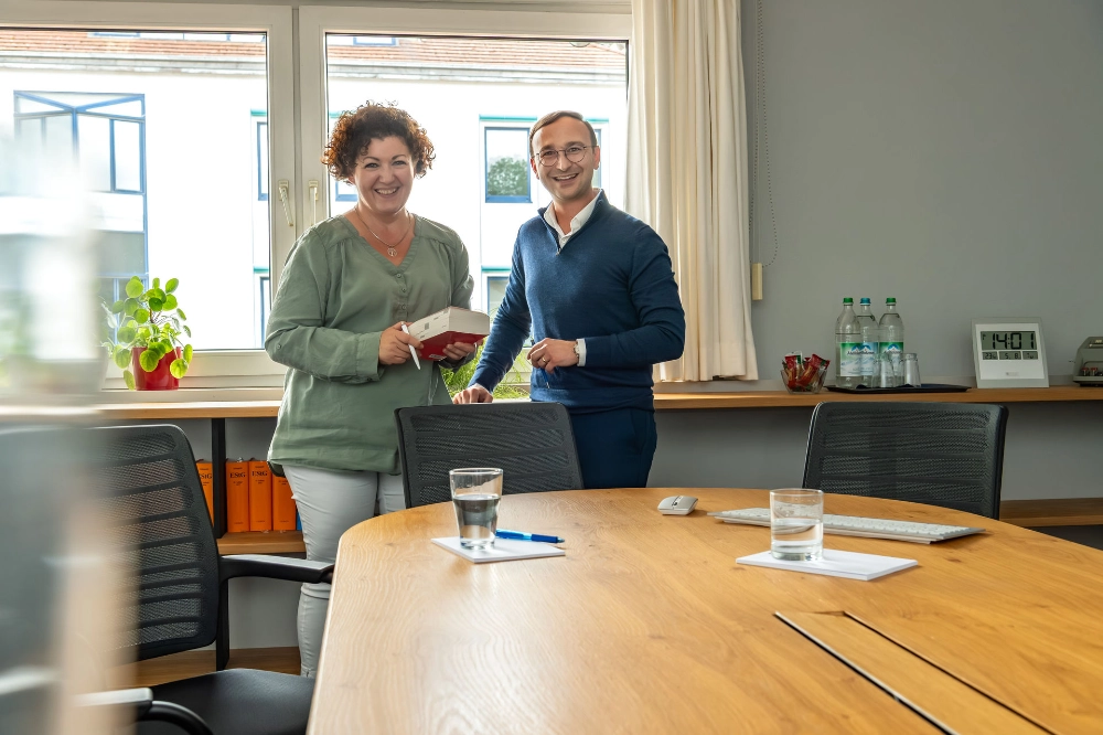 Zwei Angestellte stehen vor dem Fenster und lächeln in die Kamera